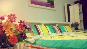 A bedroom with a bed, a bedside table, a potted plant, and a painting on the wall. - Donmuang - Impact Condo Service (Pak Kret)
