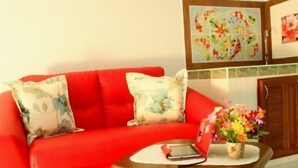 A red sofa with floral cushions, a wooden coffee table with a vase of flowers, and a framed picture on the wall. - Donmuang - Impact Condo Service (Pak Kret)