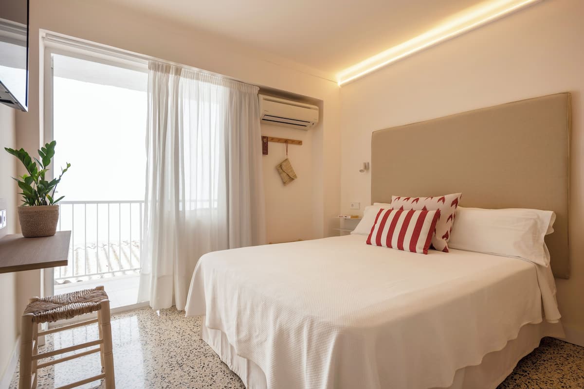 A hotel room with a bed, a desk, a chair, a potted plant, an air conditioning unit, and a view of the outside.