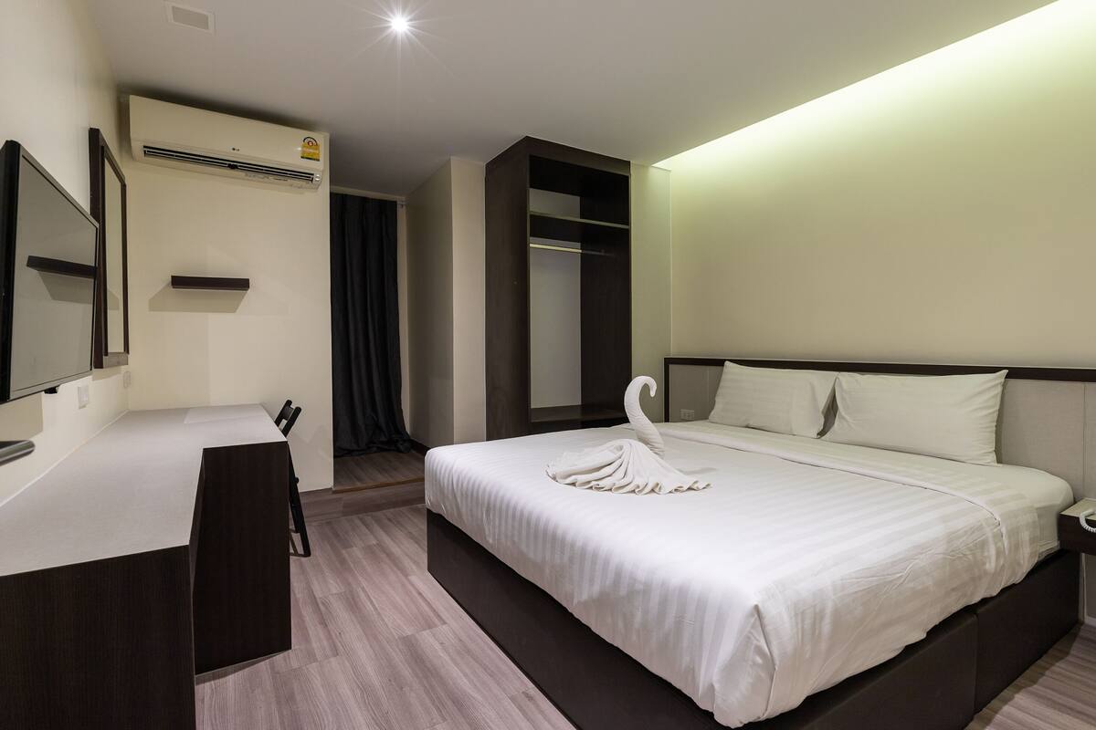 A hotel room with a bed, a desk, a mirror, a wardrobe, and an air conditioning unit.