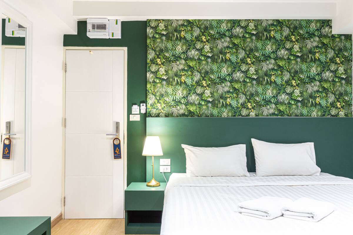 A hotel room with a green leaf-patterned wall, a white bed with two pillows, a bedside table with a lamp, and a door with a sign.
