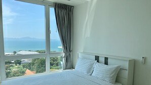 A hotel room with a large bed, a bedside table, a nightstand, and a view of the sea and mountains through a large window. - The View Cosy Beach by PSR ASIA (Pattaya)