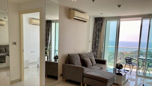 A modern living room with a grey sectional sofa, a small side table, and a view of the ocean through large windows. - The View Cosy Beach by PSR ASIA (Pattaya)