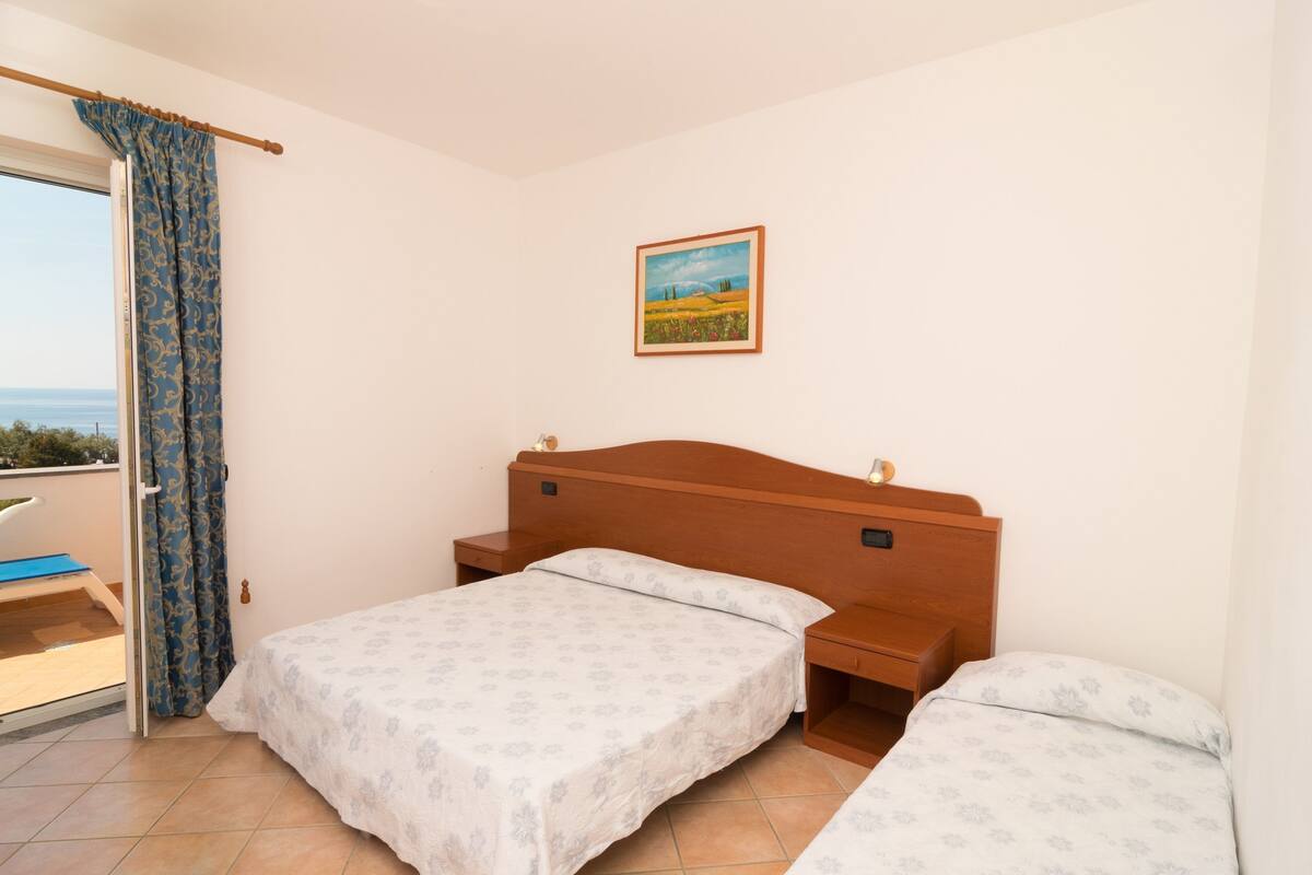 A bedroom with two beds, a wooden headboard, a painting on the wall, and a view of the sea through a window with curtains.