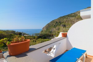 Un balcón con una silla azul, macetas de terracota con plantas y vistas al mar y a una colina.