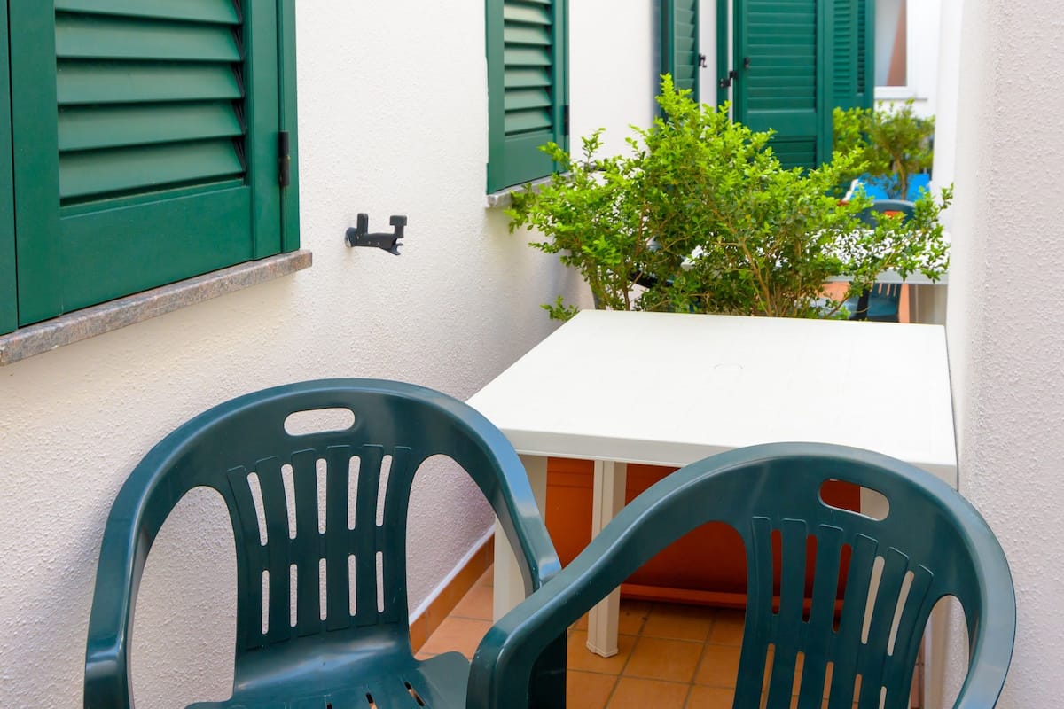 A small outdoor seating area with two green plastic chairs and a white table, set against a white wall with green shutters.