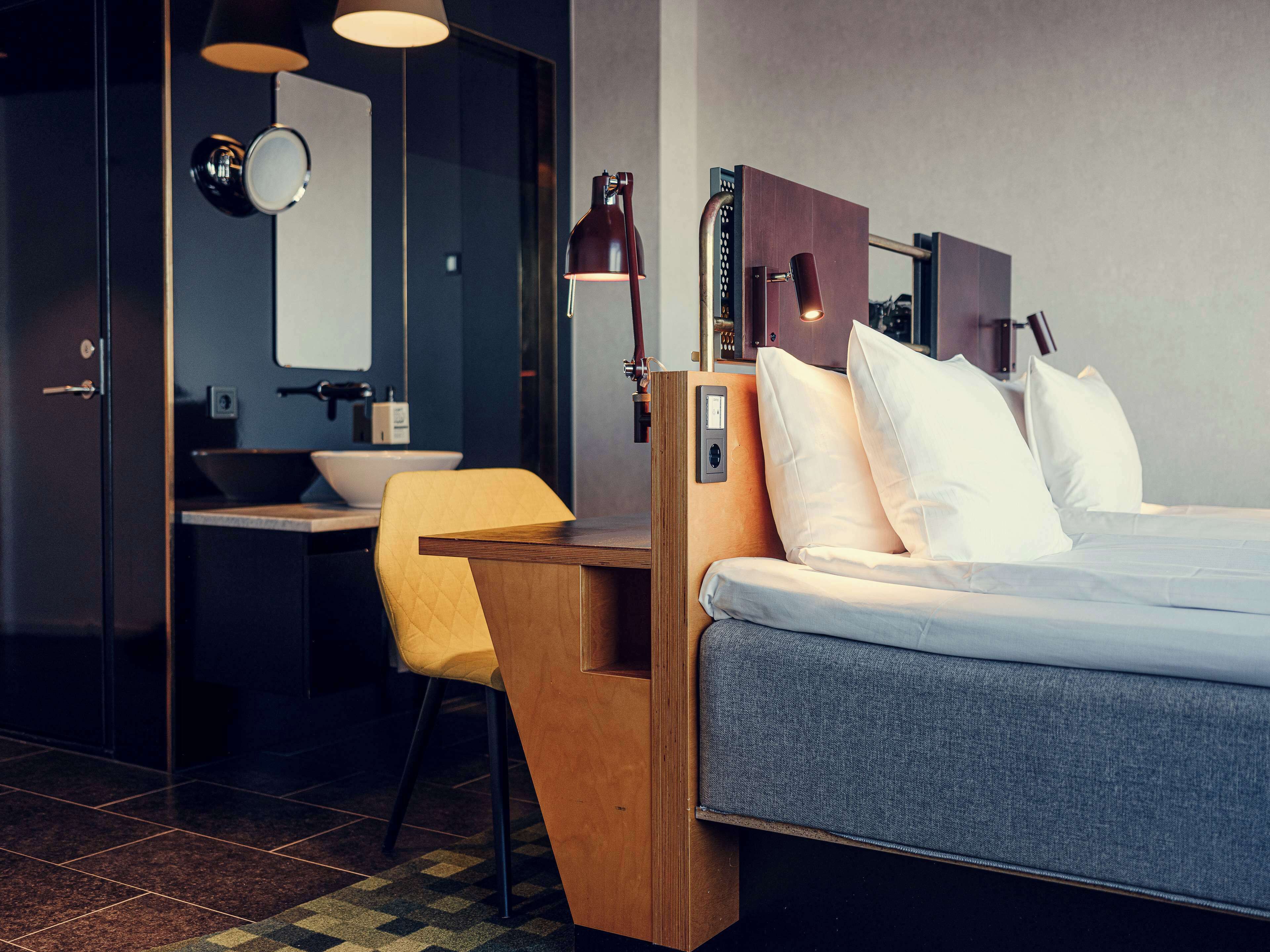 A modern hotel room with a bed, bedside table, and a small desk with a chair. There is a bathroom with a sink and mirror.