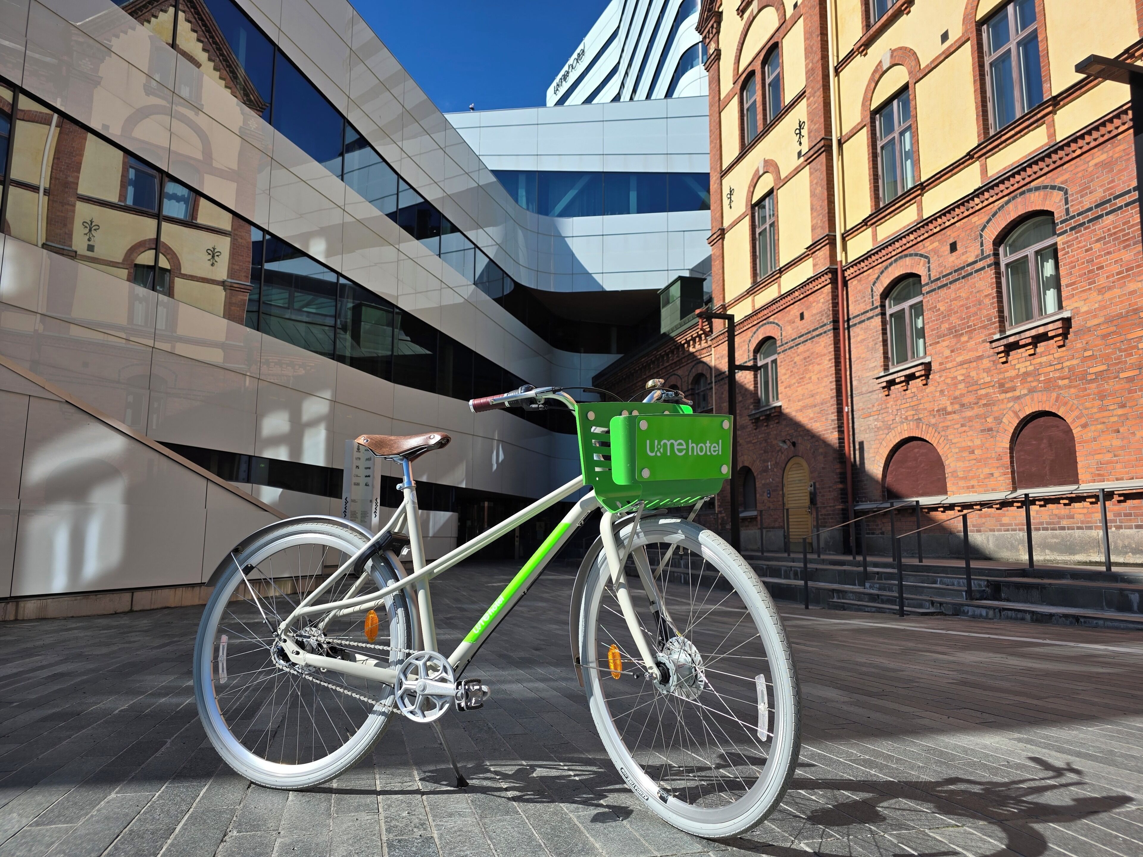 A bicycle with a green basket labeled 'Umehotel' parked in front of a modern building with a glass facade and a brick building with arched windows.