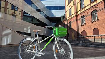 A bicycle with a green basket labeled 'Umehotel' parked in front of a modern building with a glass facade and a brick building with arched windows.