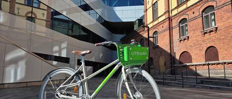 A bicycle with a green basket labeled 'Umehotel' parked in front of a modern building with a glass facade and a brick building with arched windows.