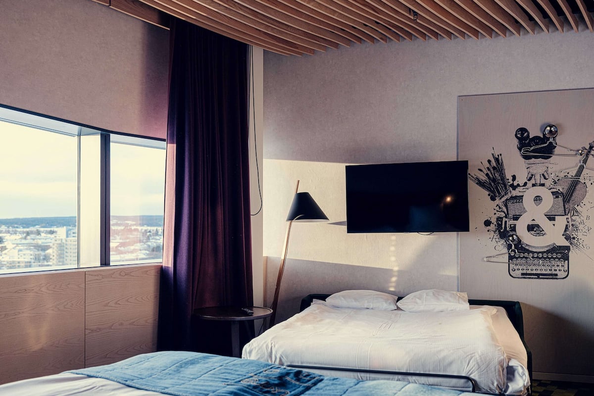 A hotel room with two beds, a large window with a view, a flat-screen TV, and a wall art piece.