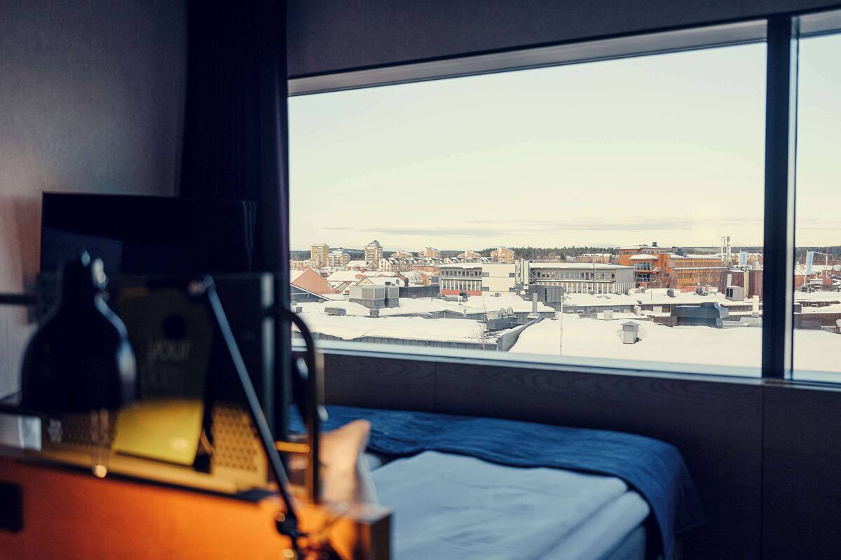 A hotel room with a bed, a desk with a laptop, and a window offering a city view.