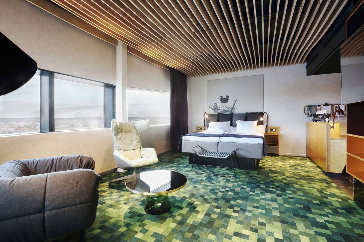 A modern hotel room with a large bed, a chair, a small table, and a view of the city through large windows.