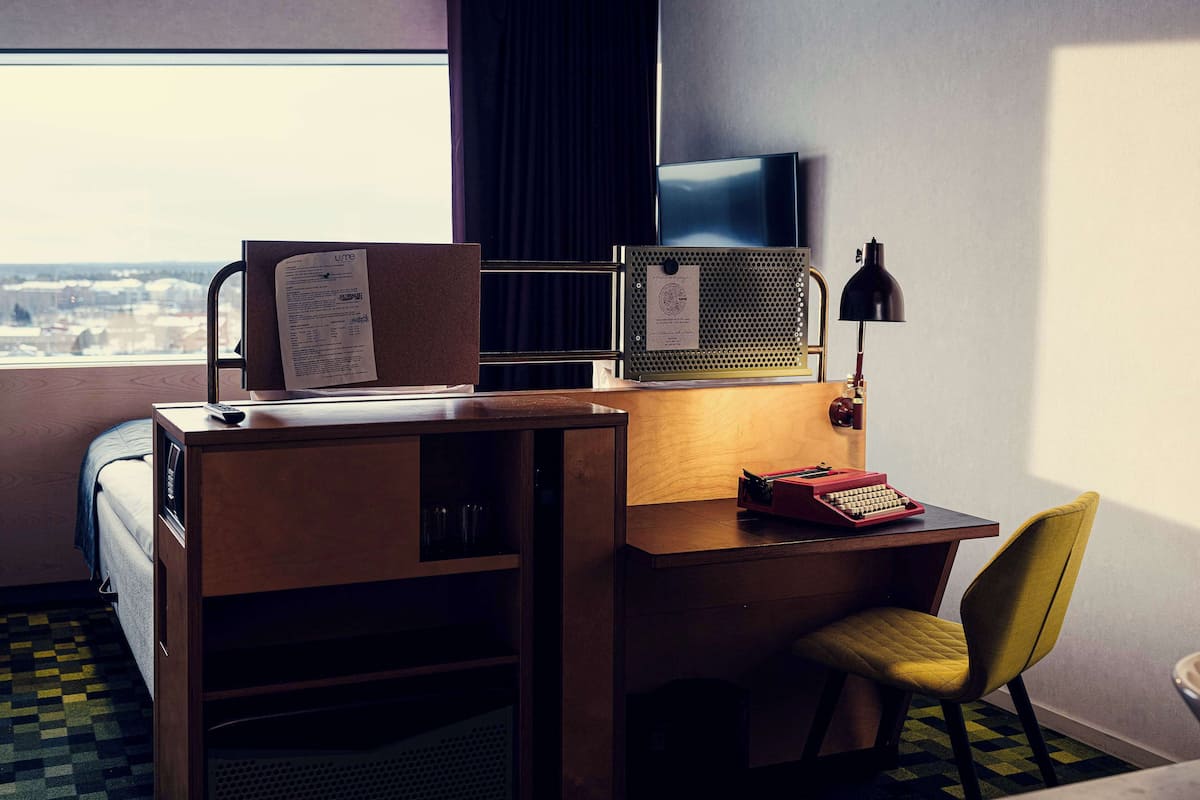 A hotel room with a desk, a chair, a TV, a lamp, and a window with a view.