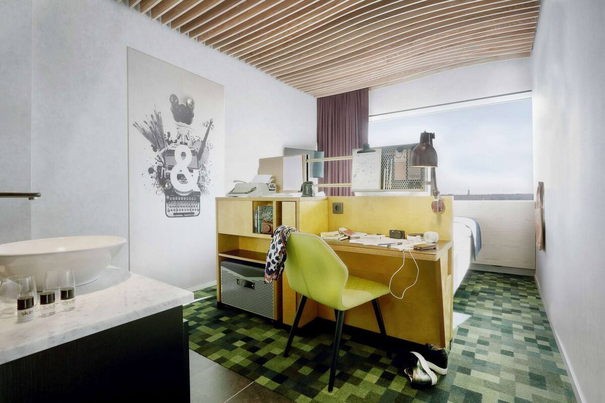A modern hotel room with a wooden ceiling, a desk with a chair, a large window, and a bathroom with a sink and mirror.