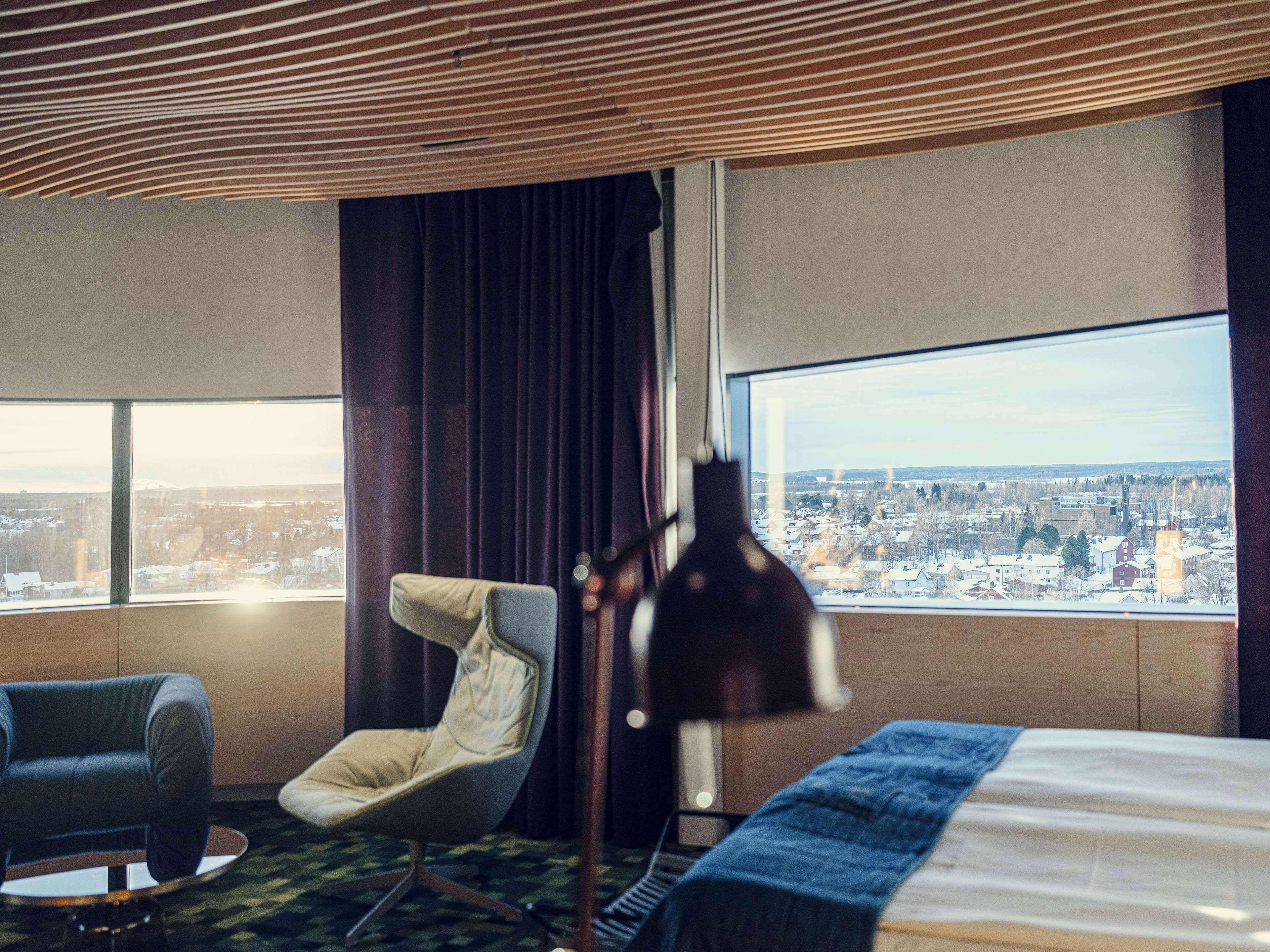 A hotel room with a large window, a bed, a chair, and a lamp.