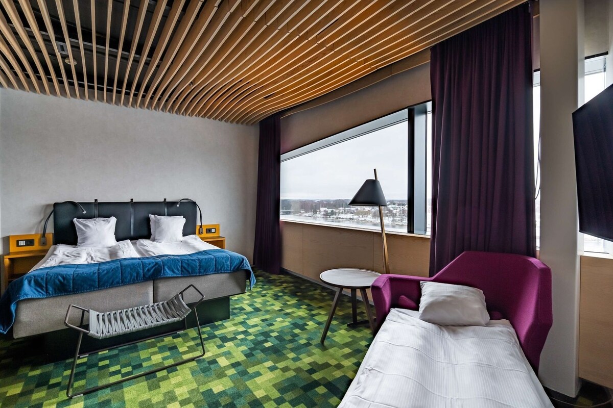A hotel room with a bed, a chair, a small table, a window with a view, and a ceiling with wooden slats.
