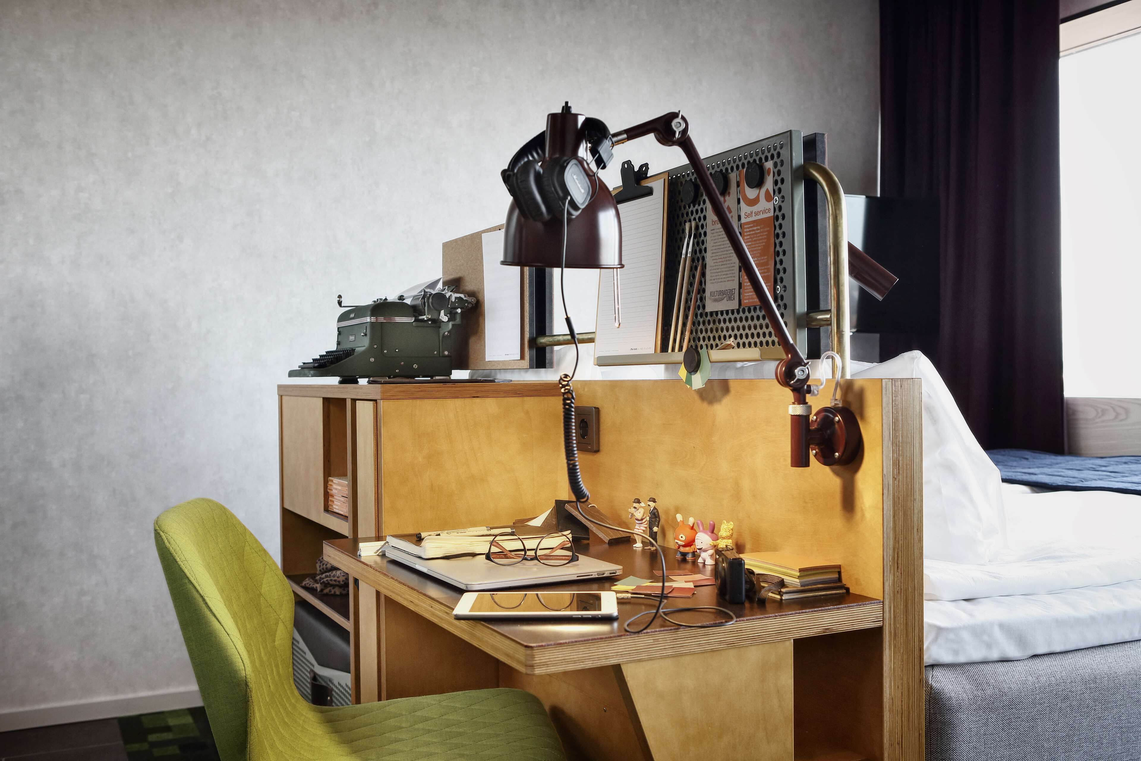 A compact hotel room with a wooden desk, a green chair, a bed with white linens, a desk lamp, and a small desk lamp with headphones.