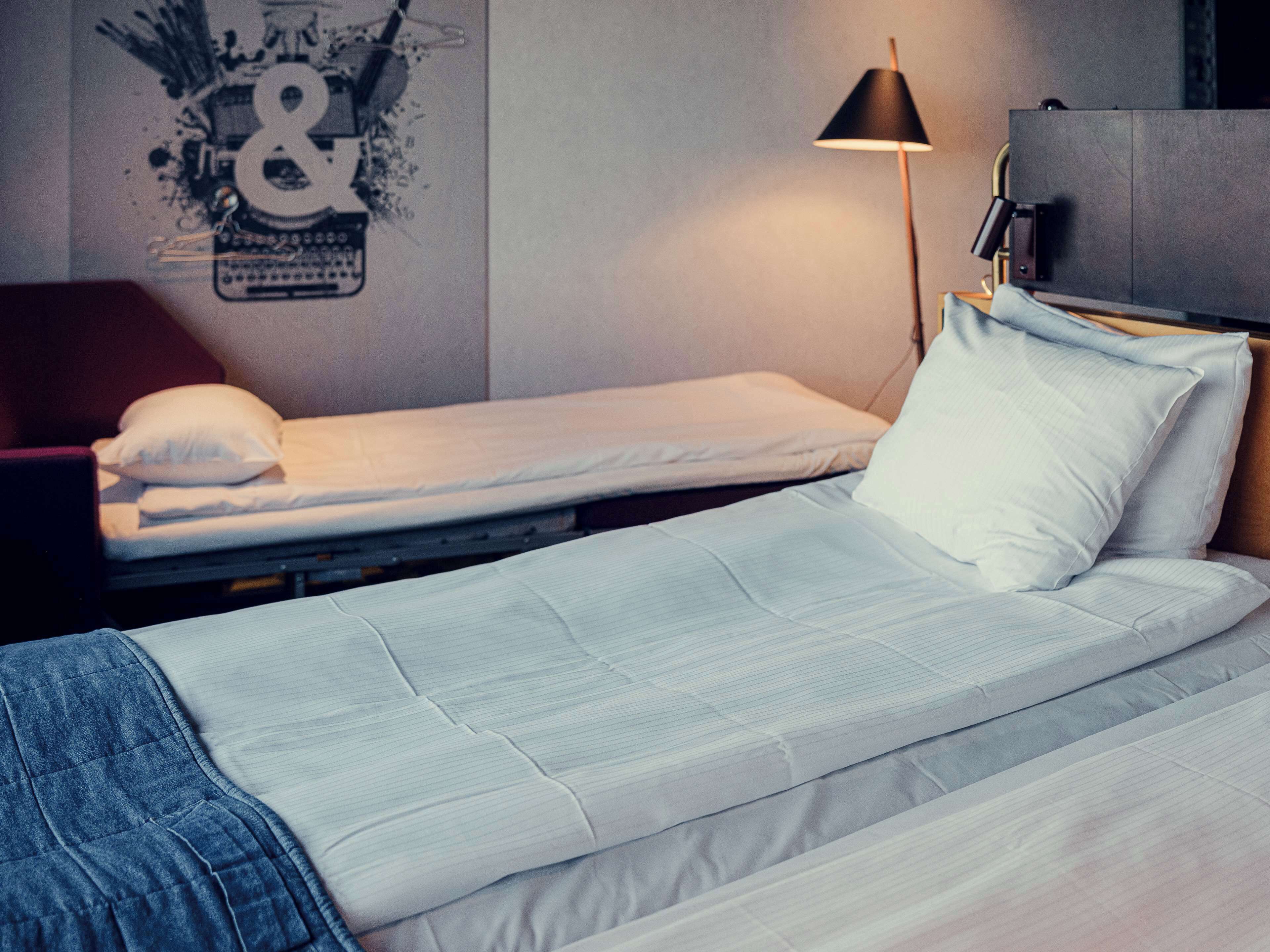 A single bed with a white mattress and pillow, a red chair, a black lamp, and a wall with a graphic.