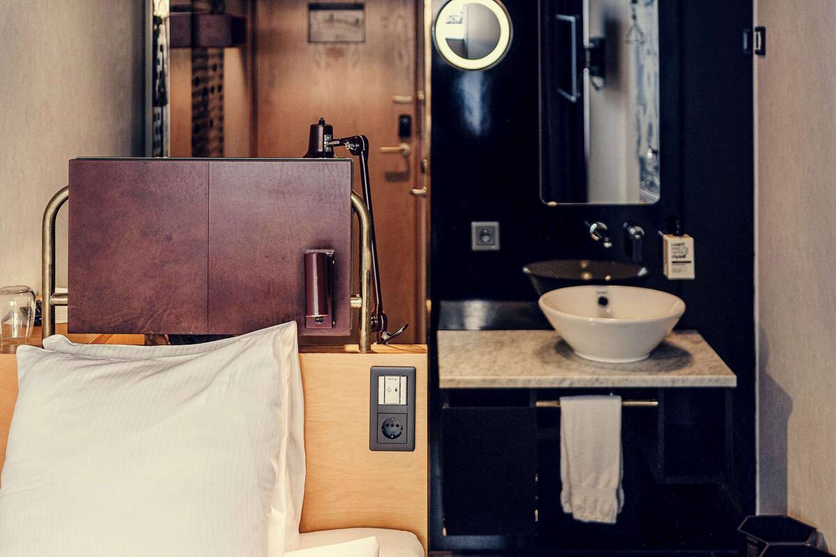 A hotel room with a bed, a bedside table, a mirror, a sink, and a towel hanging on the wall.