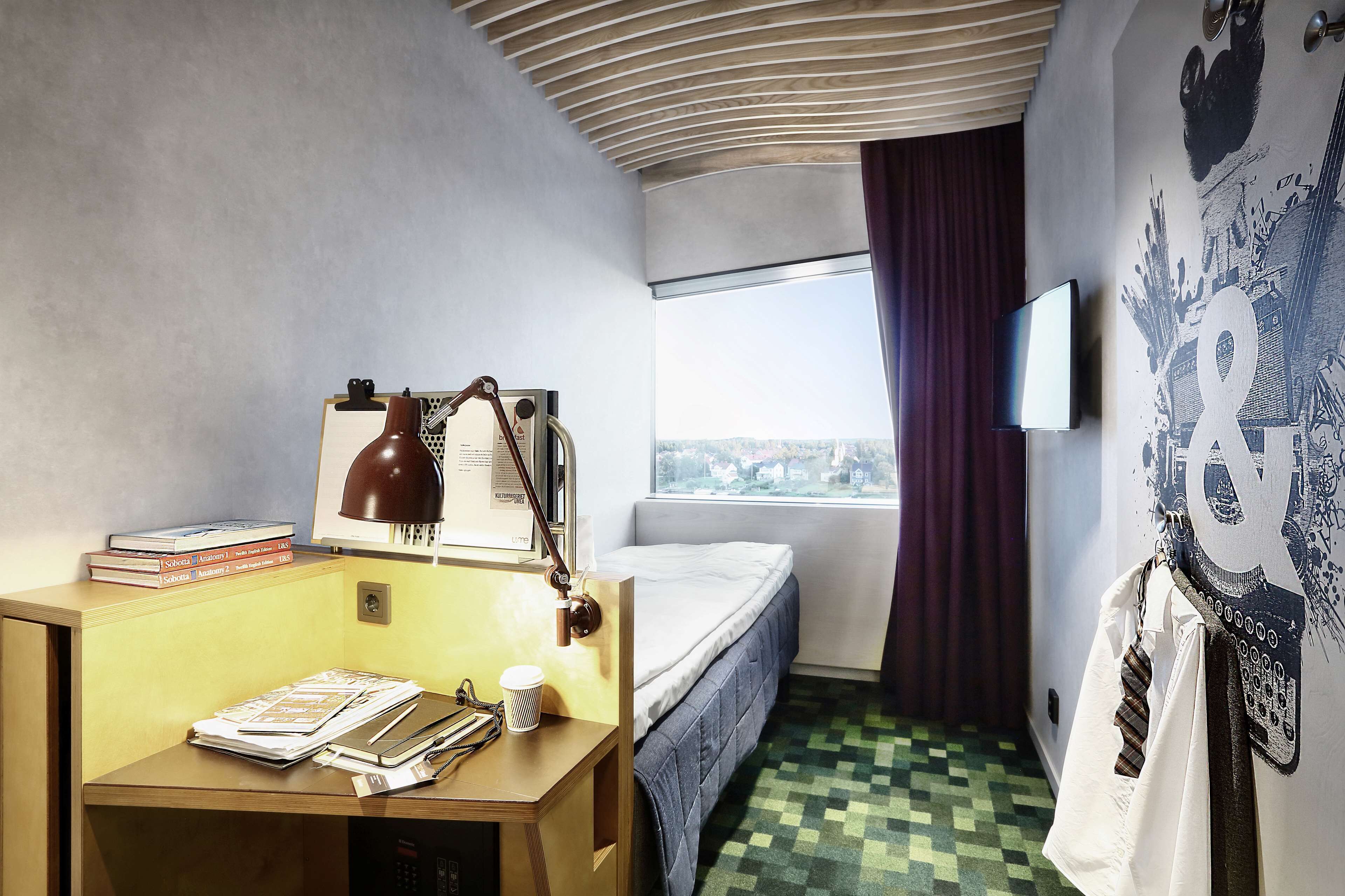 A compact hotel room with a desk, a bed, a window with a view, and a wall-mounted TV.