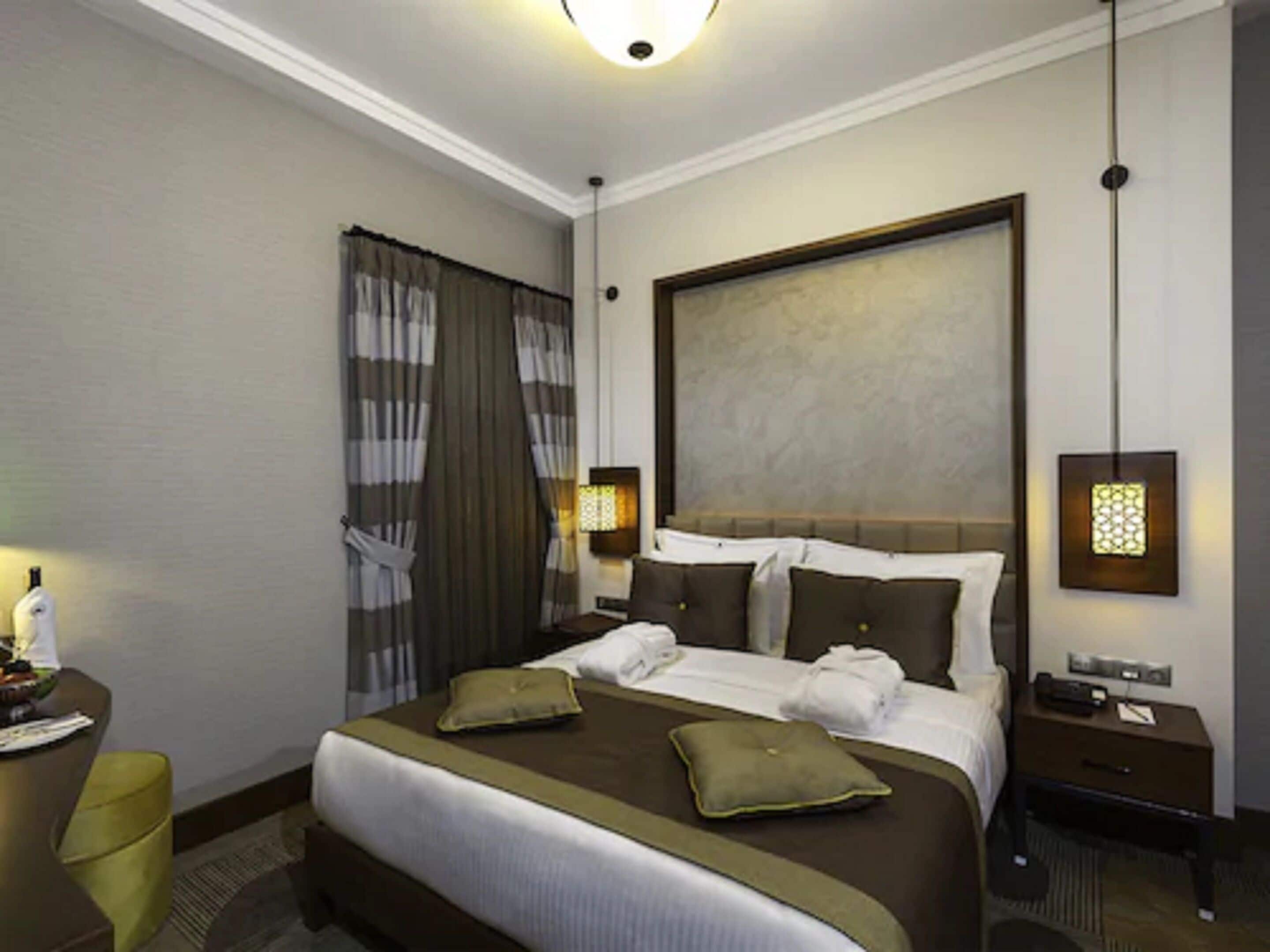 Een hotelkamer met een groot bed, nachtkastje, lamp en een raam met gordijnen.