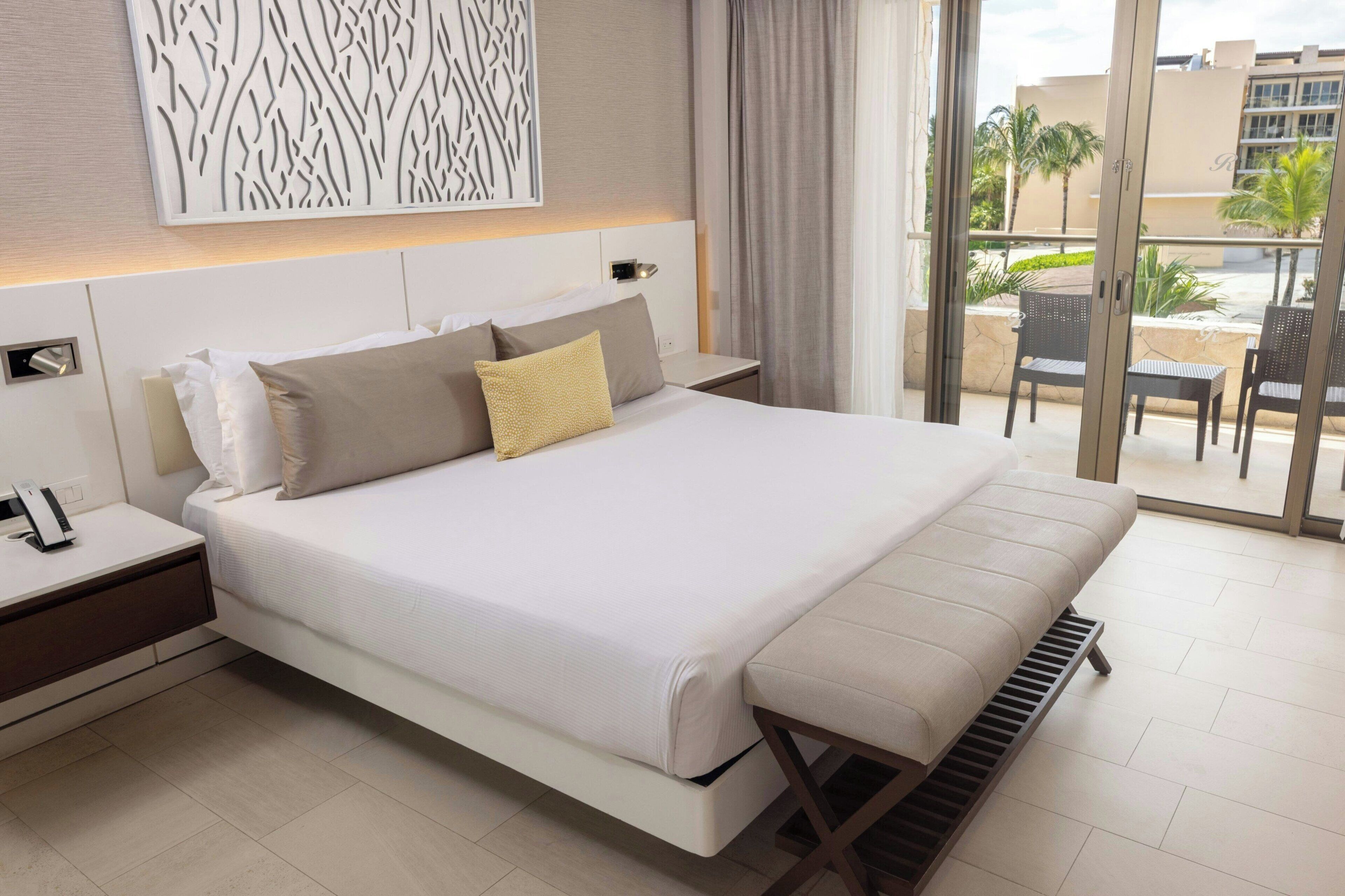 Una habitación de hotel moderna con una cama grande, una mesita de noche con teléfono, un banco y vistas al exterior.