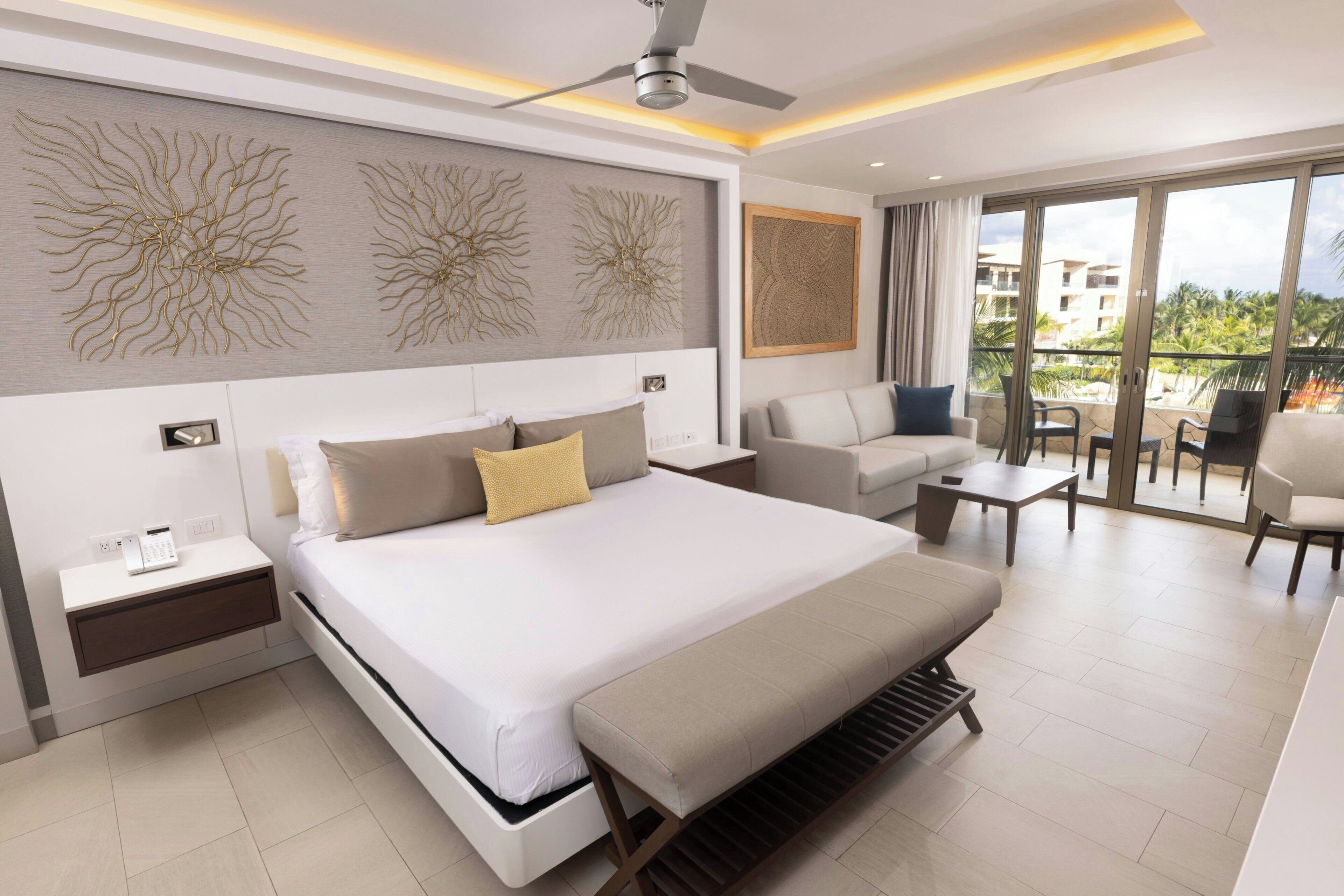A modern hotel room with a large bed, a sofa, a coffee table, and a balcony with a view of buildings and palm trees.