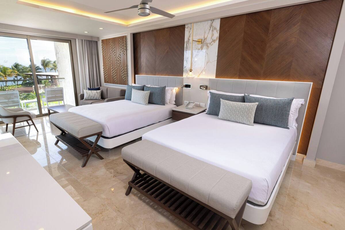 A modern hotel room with two beds, a balcony, and a view of palm trees.