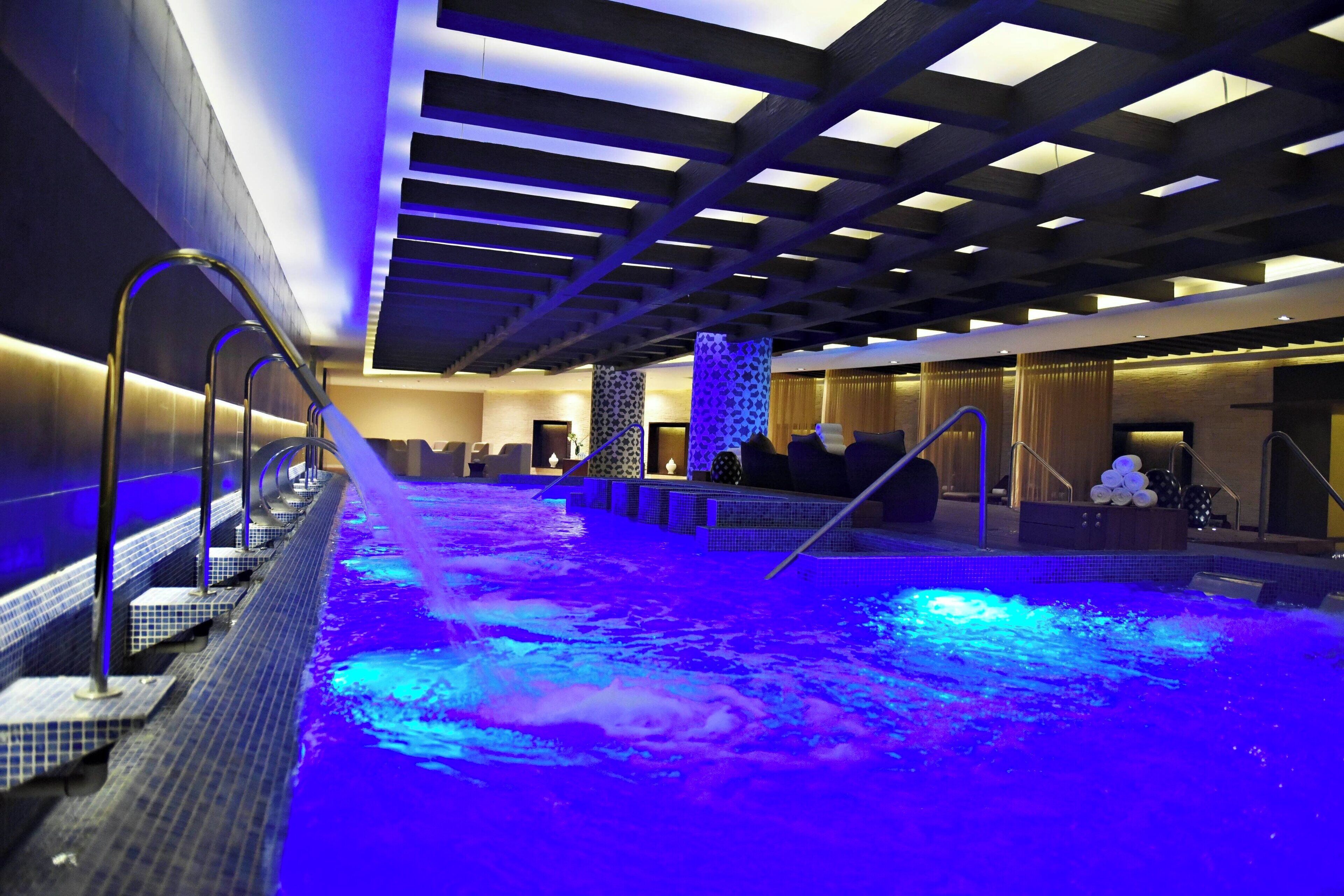 Una piscina interior moderna con un elemento acuático iluminado en azul, una zona de estar y un mostrador de recepción.