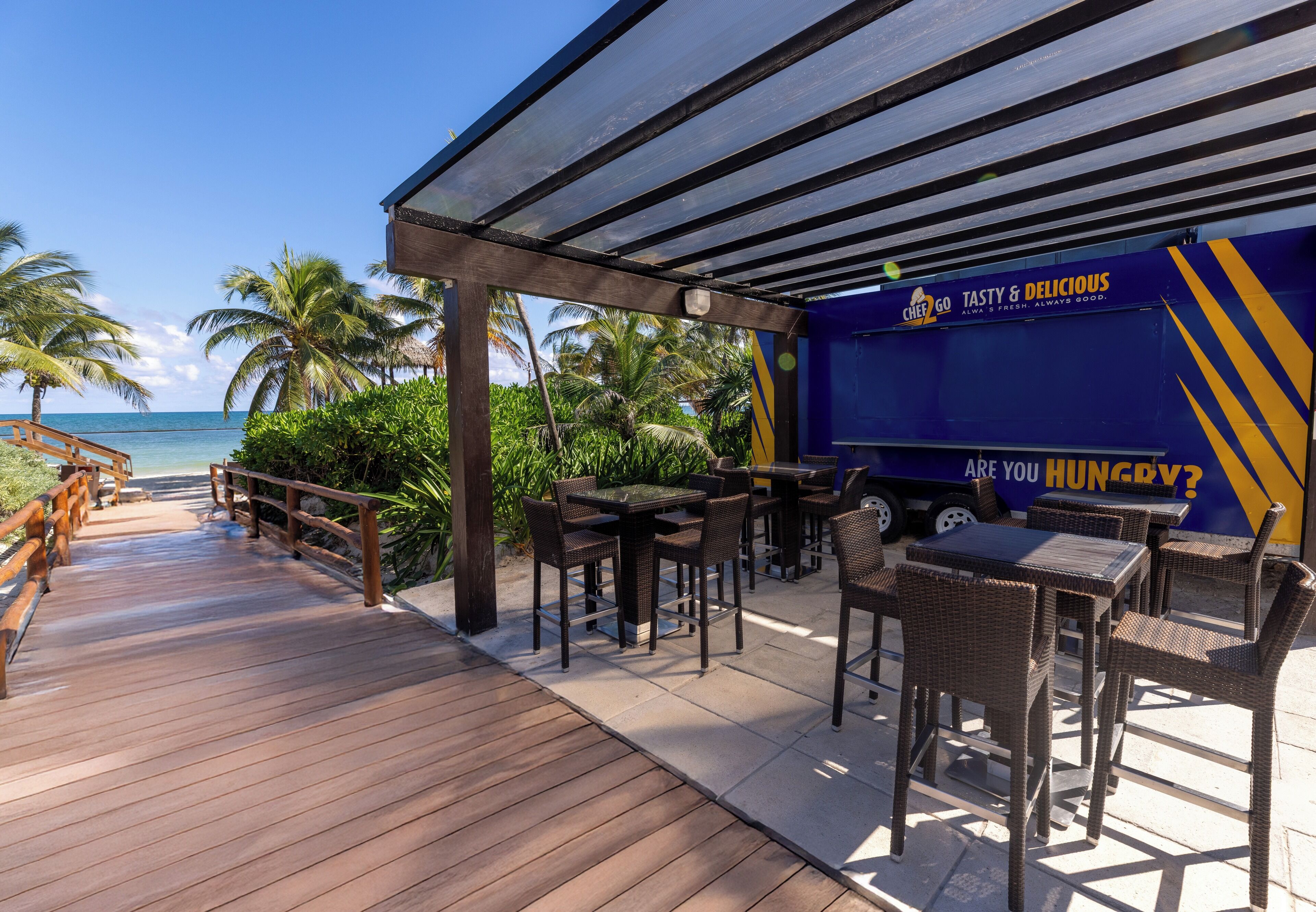 A beachside patio with wooden deck, outdoor seating, and a food truck with "Tasty & Delicious" branding.