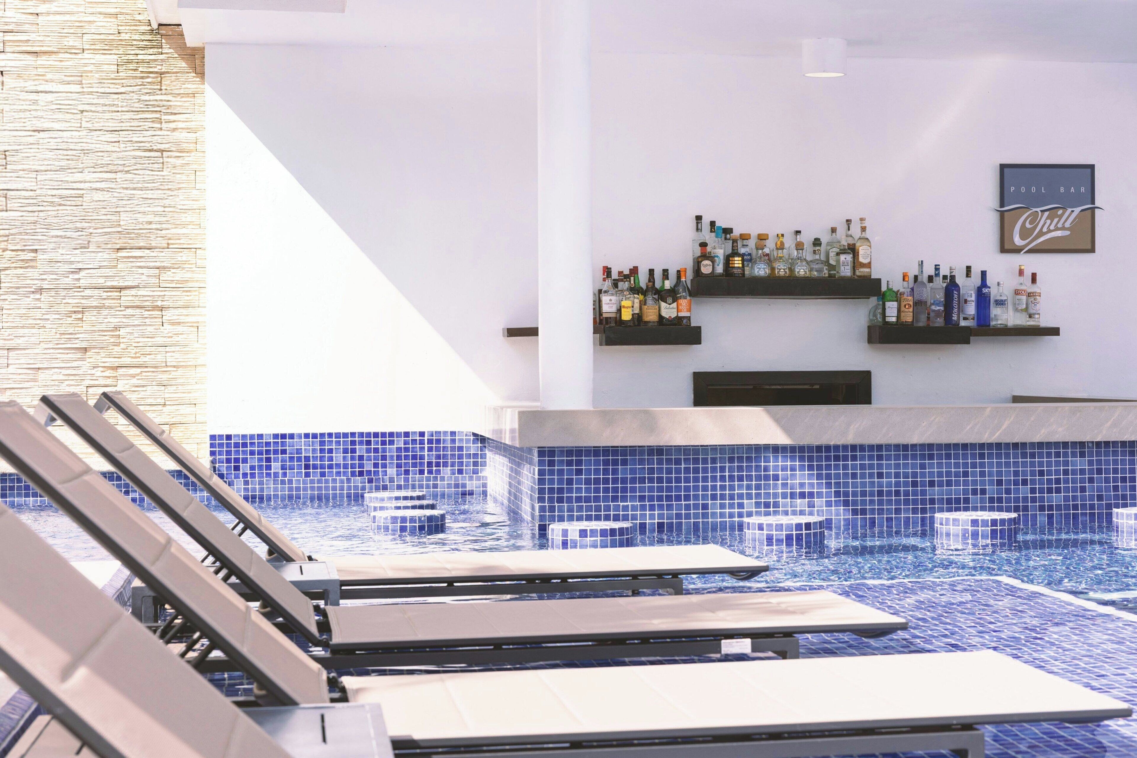 Un espace piscine avec des chaises longues, un bar doté d’étagères remplies de bouteilles et un panneau indiquant : « Bar de la piscine – Ambiance détendue ».