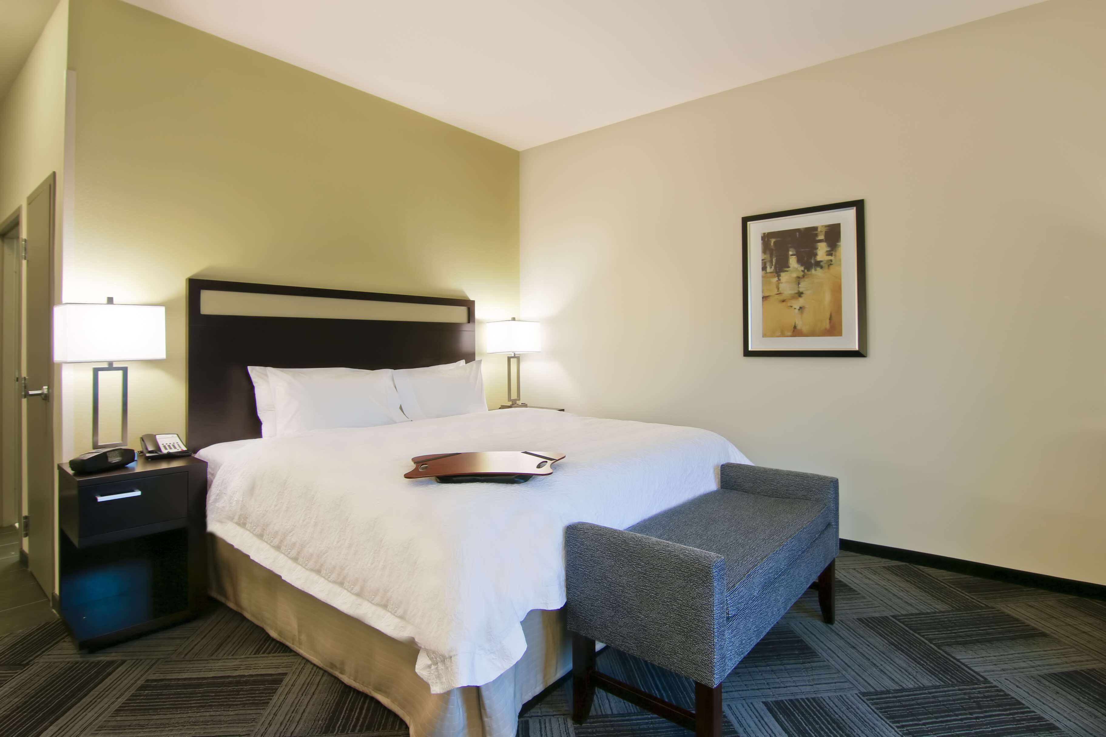 A hotel room with a bed, two bedside lamps, a nightstand with a phone, a chair, and a framed picture on the wall.