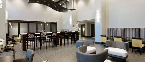 A spacious lobby with modern furniture, a high ceiling with patterned design, and a dining area with wooden chairs and tables.