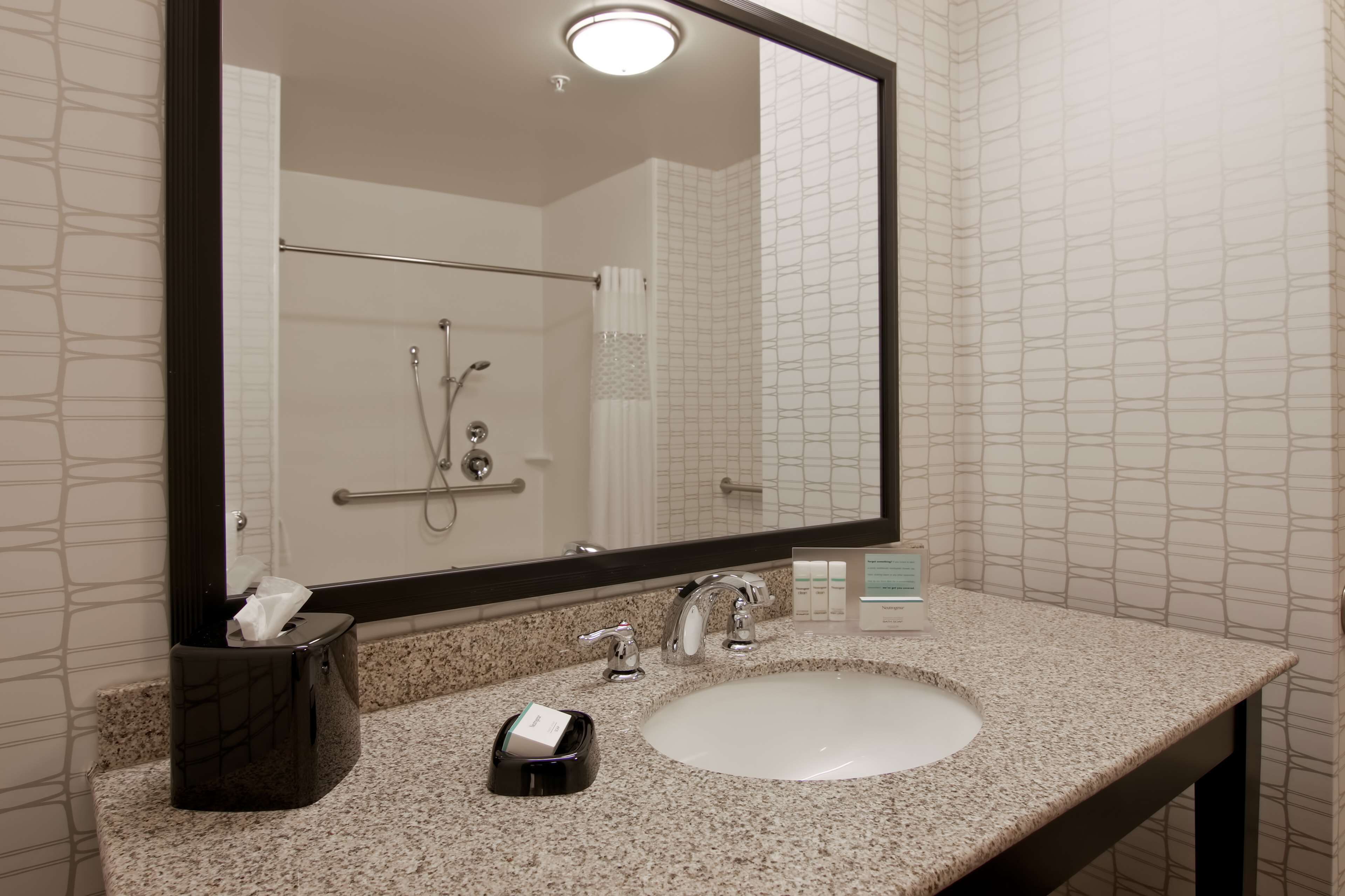 A bathroom with a large mirror, a sink, a towel dispenser, and a shower area.