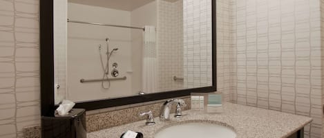 A bathroom with a large mirror, a sink, a towel dispenser, and a shower area.