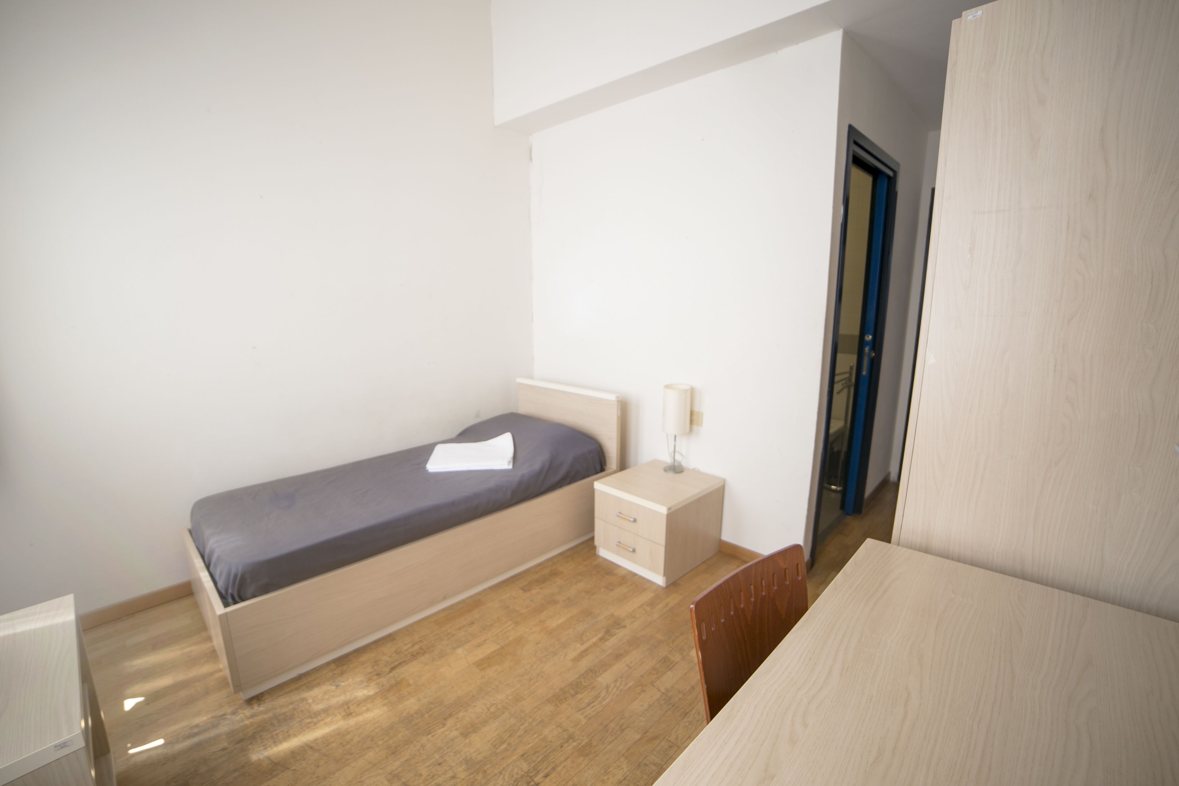 A small, single-bed room with a wooden floor, a bedside table with a lamp, and a door to the right.