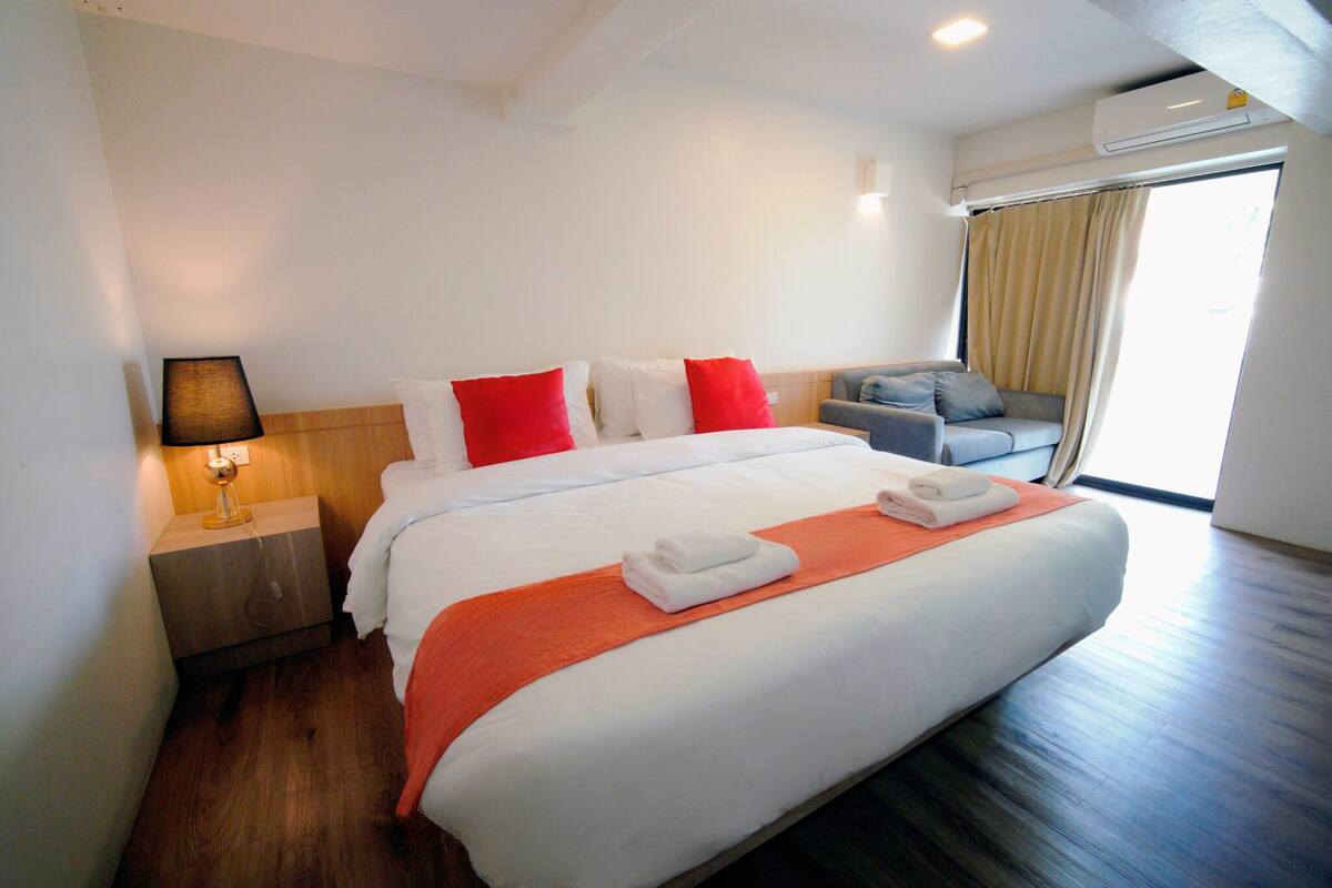 A hotel room with a large bed, a nightstand with a lamp, a sofa, and a window with curtains.