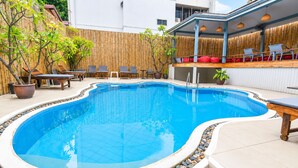 Ein Schwimmbecken mit geschwungener Kante, umgeben von einer Holzterrasse, Blumentöpfen und Gartenmöbeln.