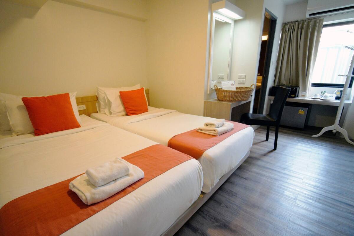 A hotel room with two beds, each with orange and white bedding, a desk with a chair, a mirror, and a window with curtains.