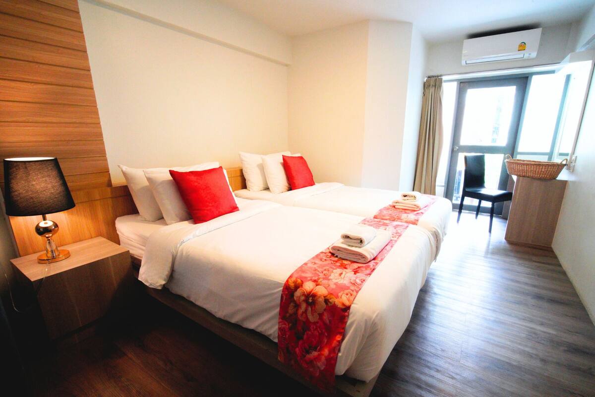 A hotel room with two beds, a nightstand with a lamp, a wooden headboard, a window with curtains, and a basket on a side table.