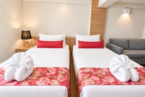 A hotel room with two beds, each with a red floral bedspread and white pillows. There is a nightstand with a lamp and a small table with a sign. A gray sofa is visible in the background.
