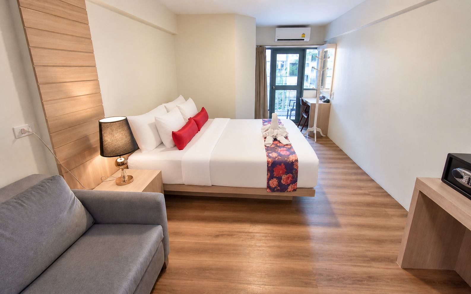 A hotel room with a bed, a bedside table with a lamp, a sofa, and a wooden floor.