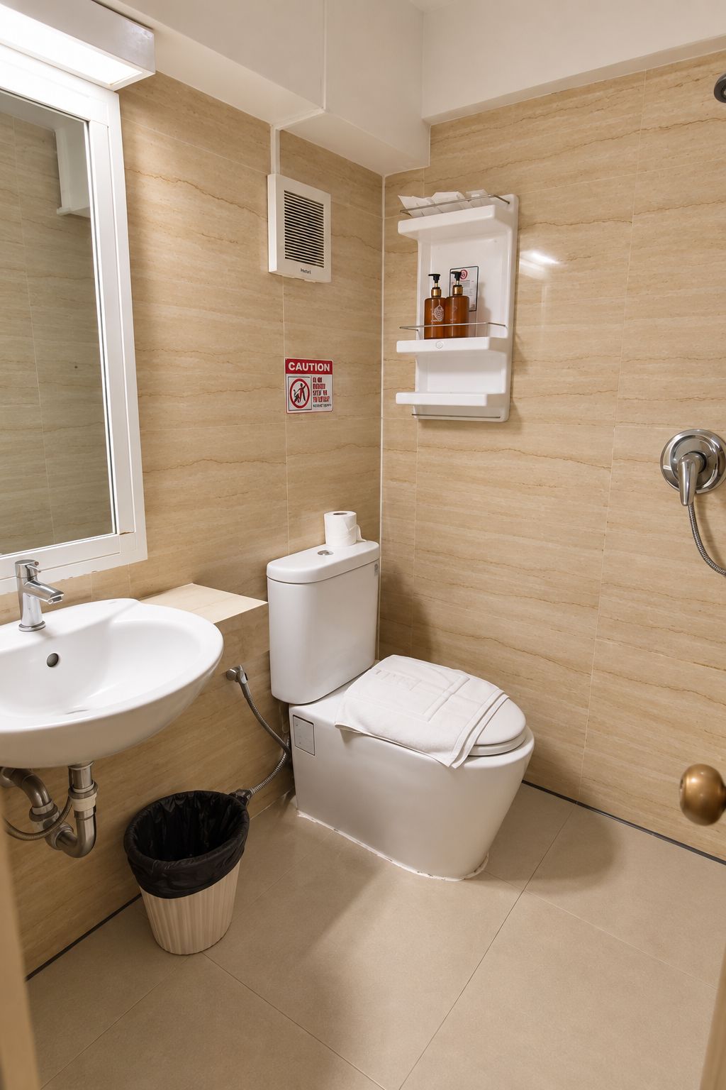 A bathroom with a sink, toilet, and shower. There is a caution sign on the wall and a trash bin.