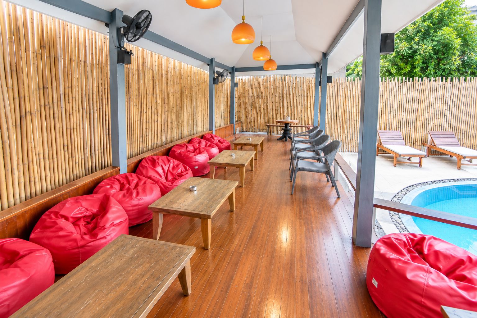 A covered outdoor seating area with wooden tables and red bean bags, a pool, and a bamboo fence.
