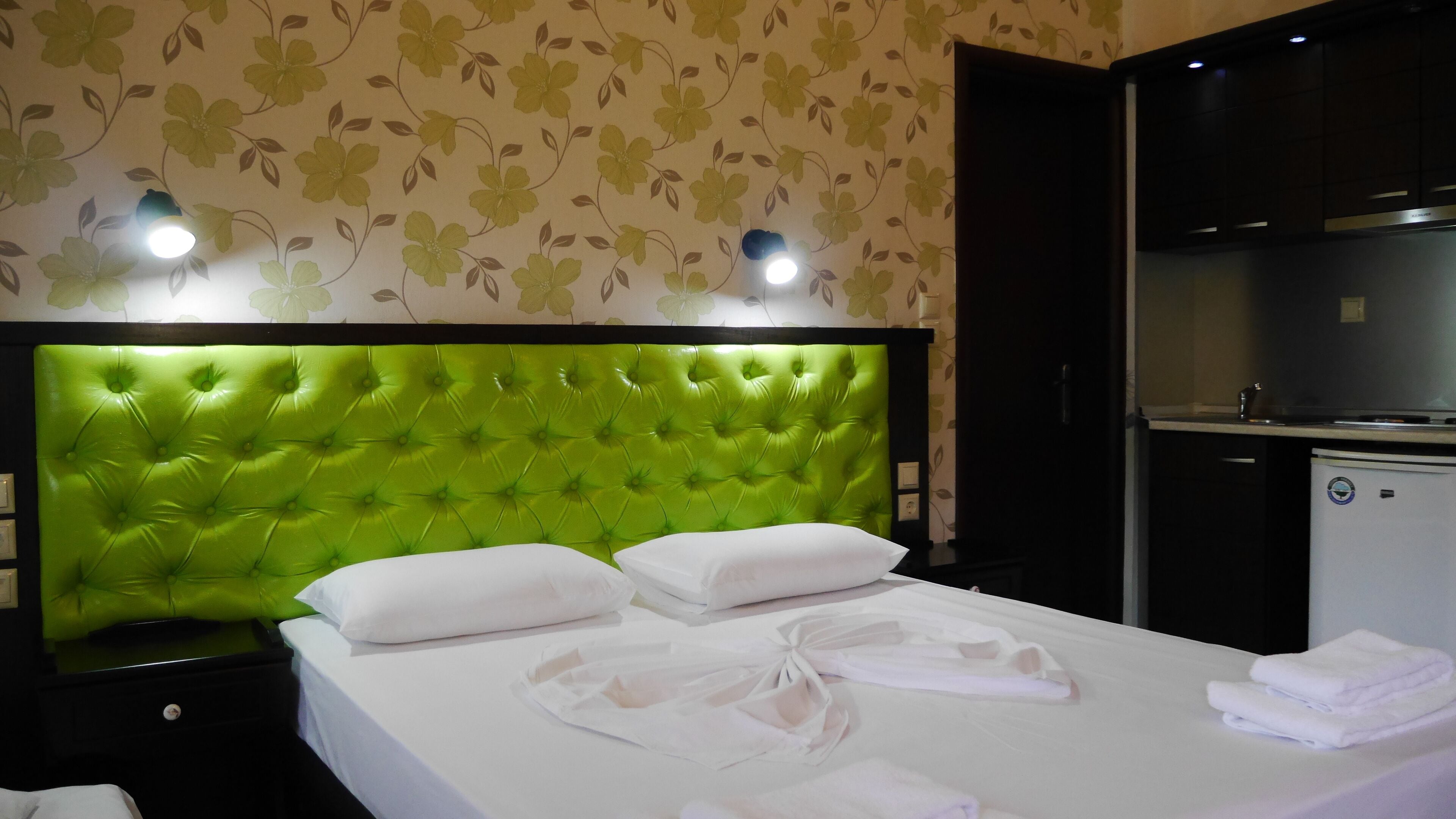A hotel room with a green tufted headboard, white bedding, and a small kitchenette.