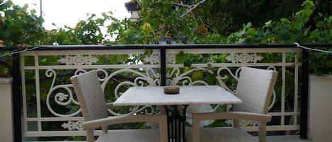 A balcony with a white table and two wicker chairs, surrounded by a decorative metal railing and lush green foliage.