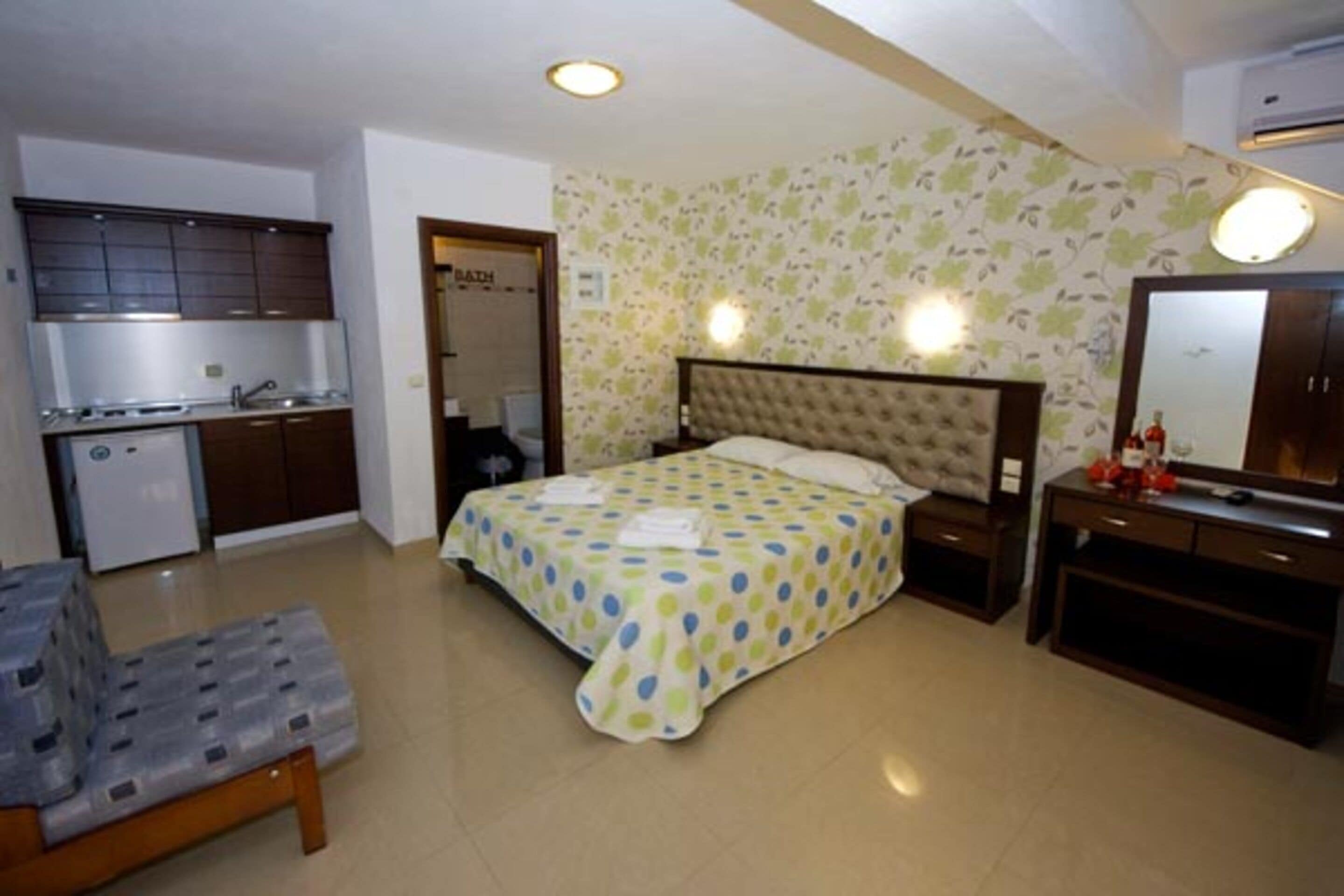 Un dormitorio con cama, mesita de noche y un pequeño escritorio con espejo. Hay una kitchenette con estufa y pileta.