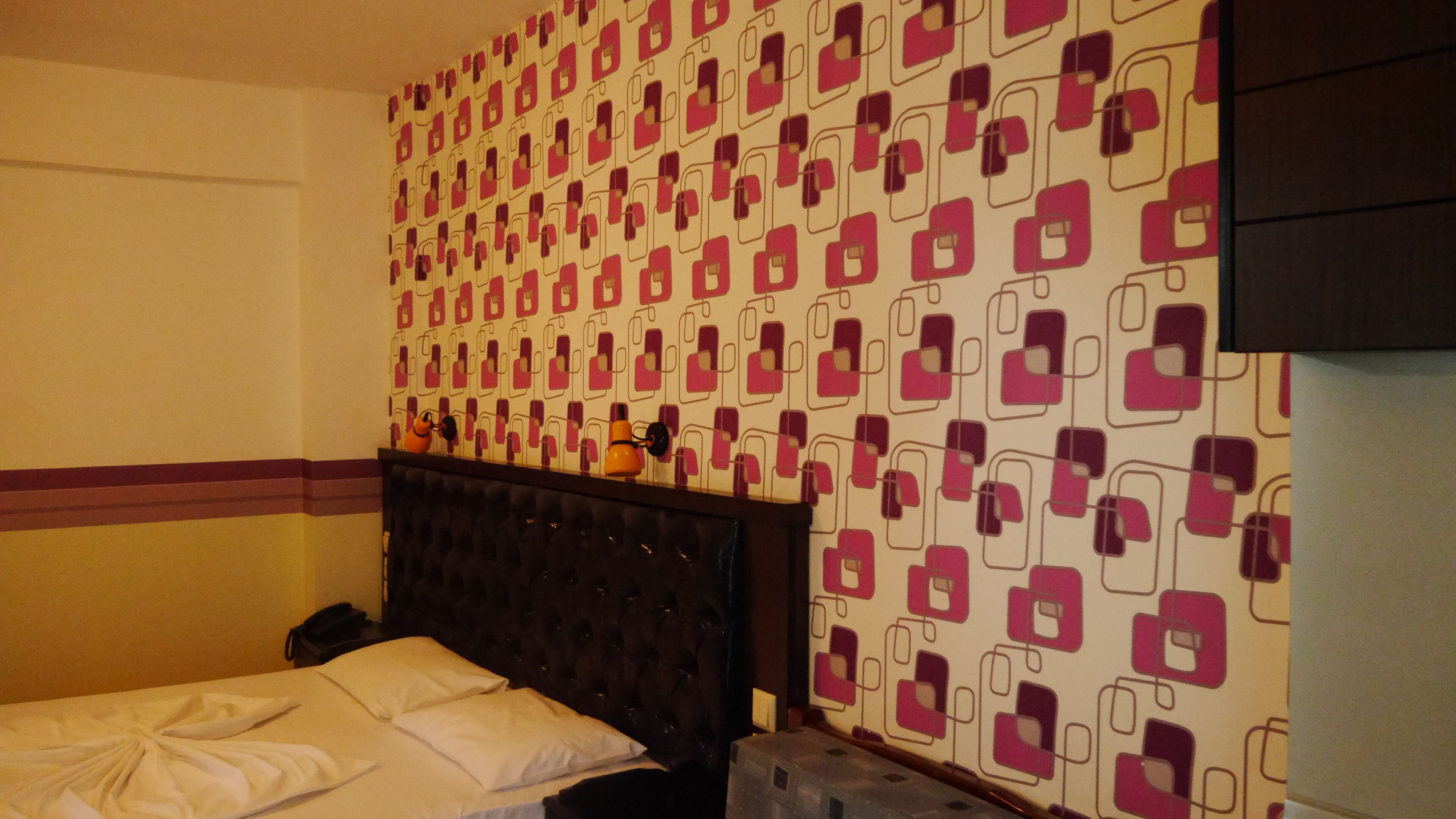 A hotel room with a bed, a headboard, a patterned wall, and a small table with a telephone.
