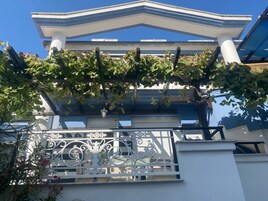 Un edificio con fachada blanca, un balcón con barandilla de hierro forjado y una pérgola cubierta de hiedra.